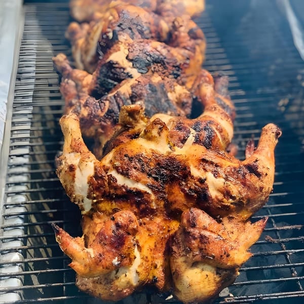 Rows of whole, seasoned chickens with crispy skin grilling on a barbecue, with smoke rising.