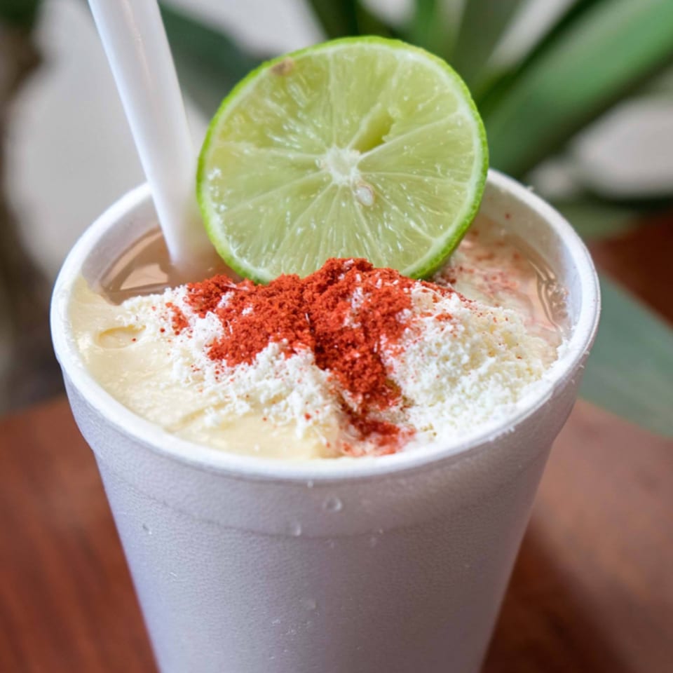Mexican esquites in a white cup, topped with white cheese, red chili powder, and a lime.