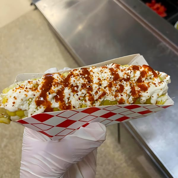 A gloved hand holds a Mexican elote, a corn on the cob covered in white cheese and red sauce.
