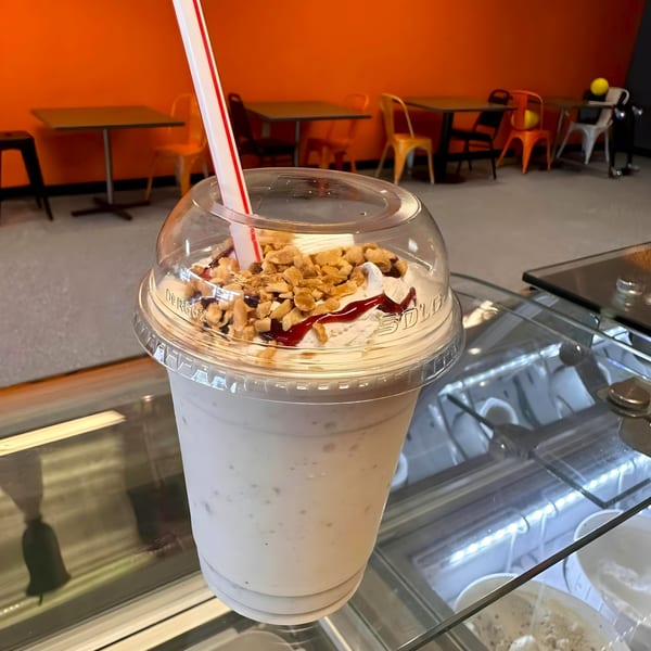 A milkshake with whipped cream, nuts, and syrup in a plastic cup, with a restaurant interior in the background.
