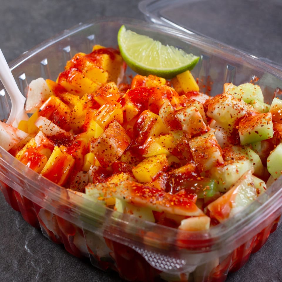A plastic container filled with diced mango, cucumber, and jicama, topped with chili powder and a lime wedge.