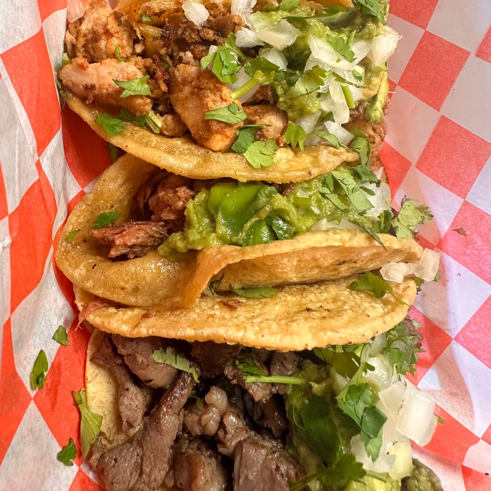 Three El Pollito Pio tacos with various fillings, guacamole, onions, and cilantro in a red-checkered basket.
