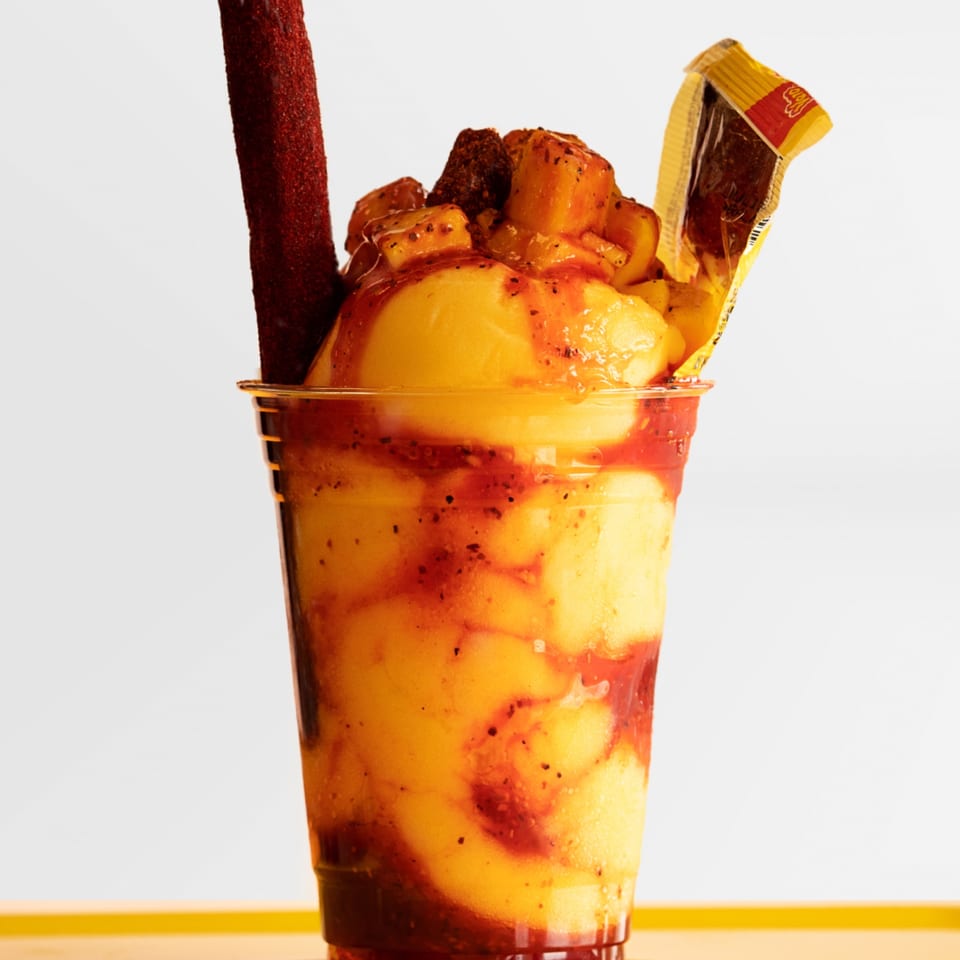 A vibrant Mexican mangonada drink in a clear cup, topped with mango chunks and a tamarind candy straw.